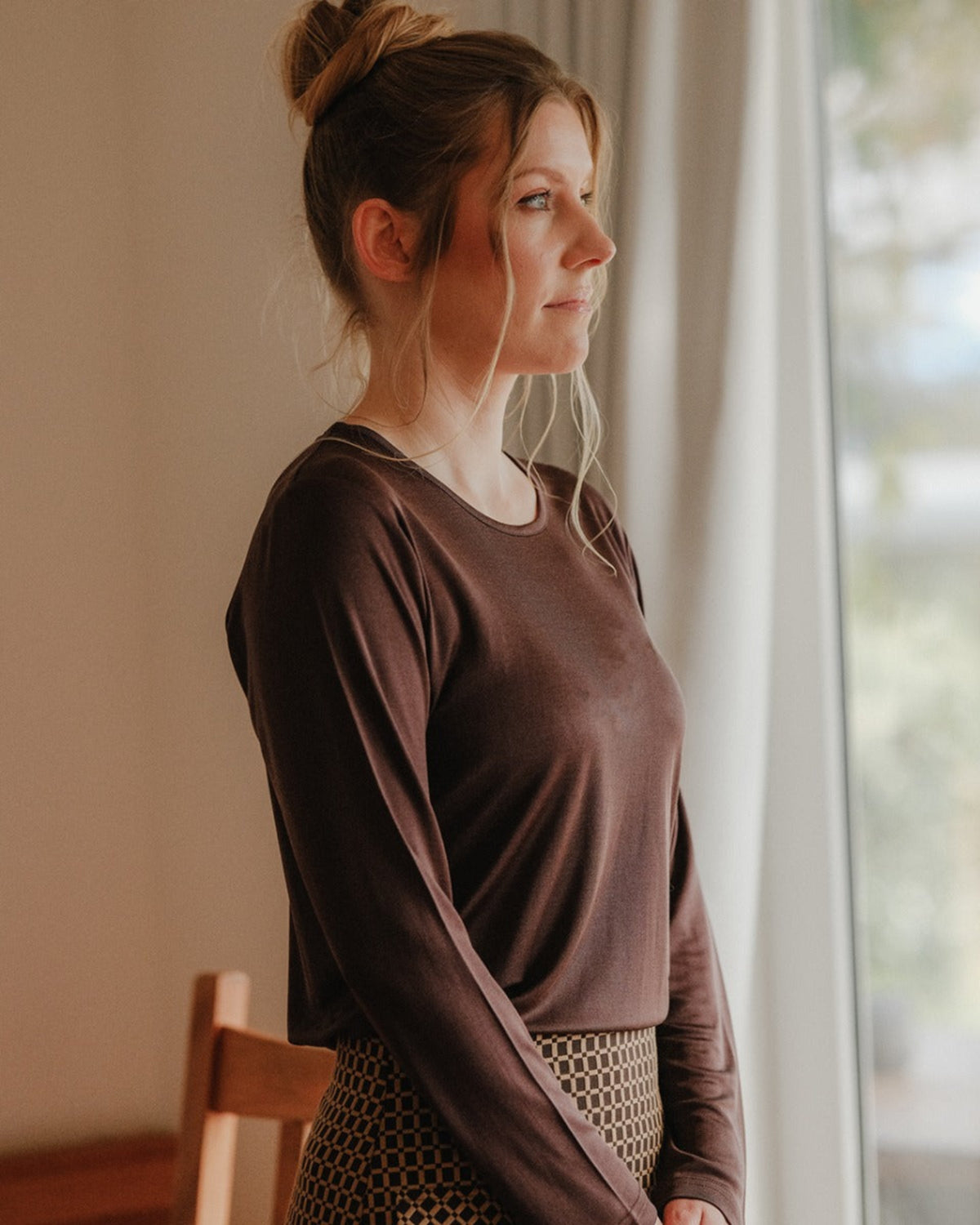 Woman in a silk brown long-sleeve top standing by a window indoors