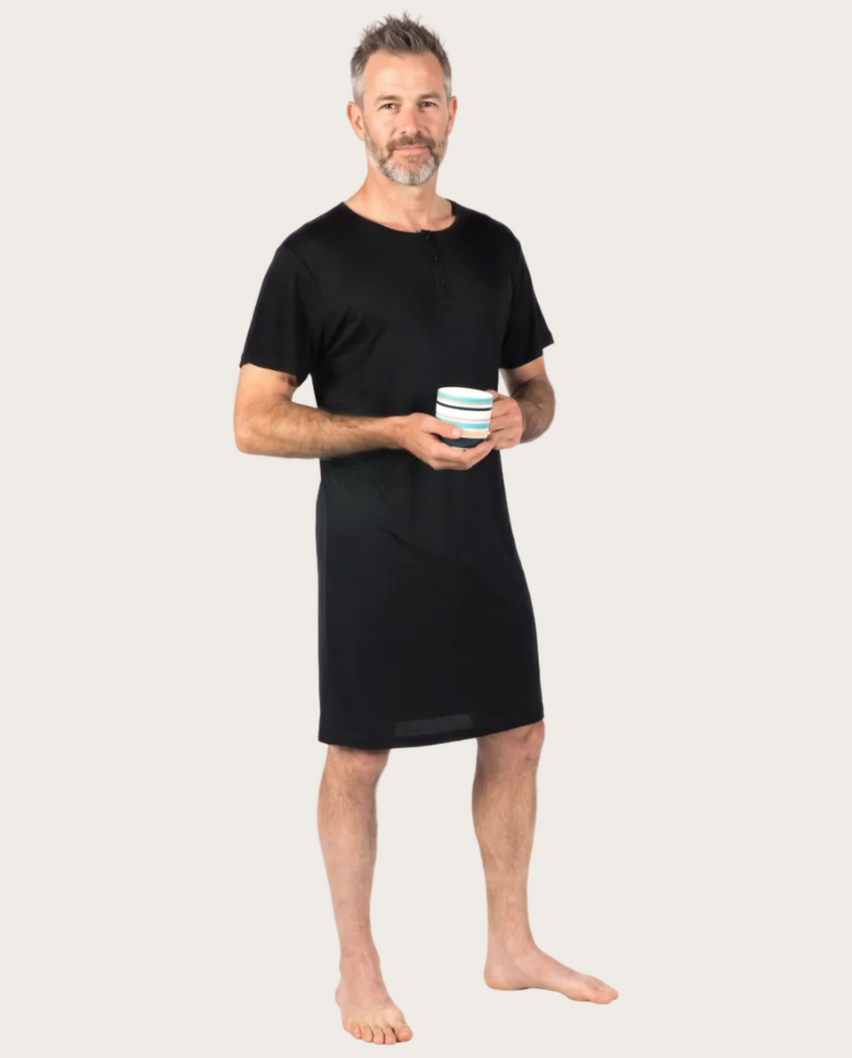 A man wearing a black short-sleeve nightshirt or tunic dress, holding a striped mug, standing against a plain white background.