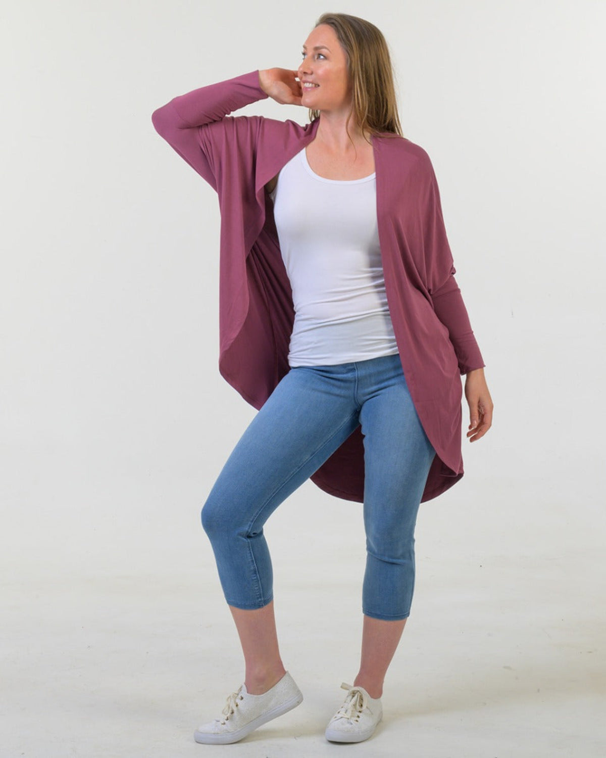 Woman wearing a bamboo cocoon mauve cardigan, white shirt, blue jeans, and white sneakers on a white background
