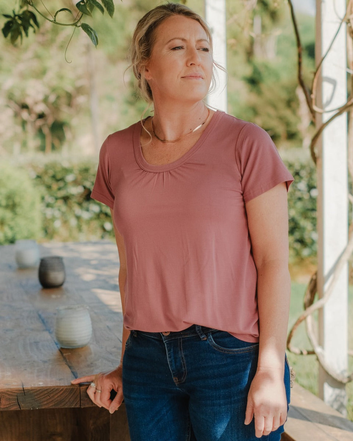 Woman wearing a pink shirt and blue jeans standing outdoors with greenery in the background