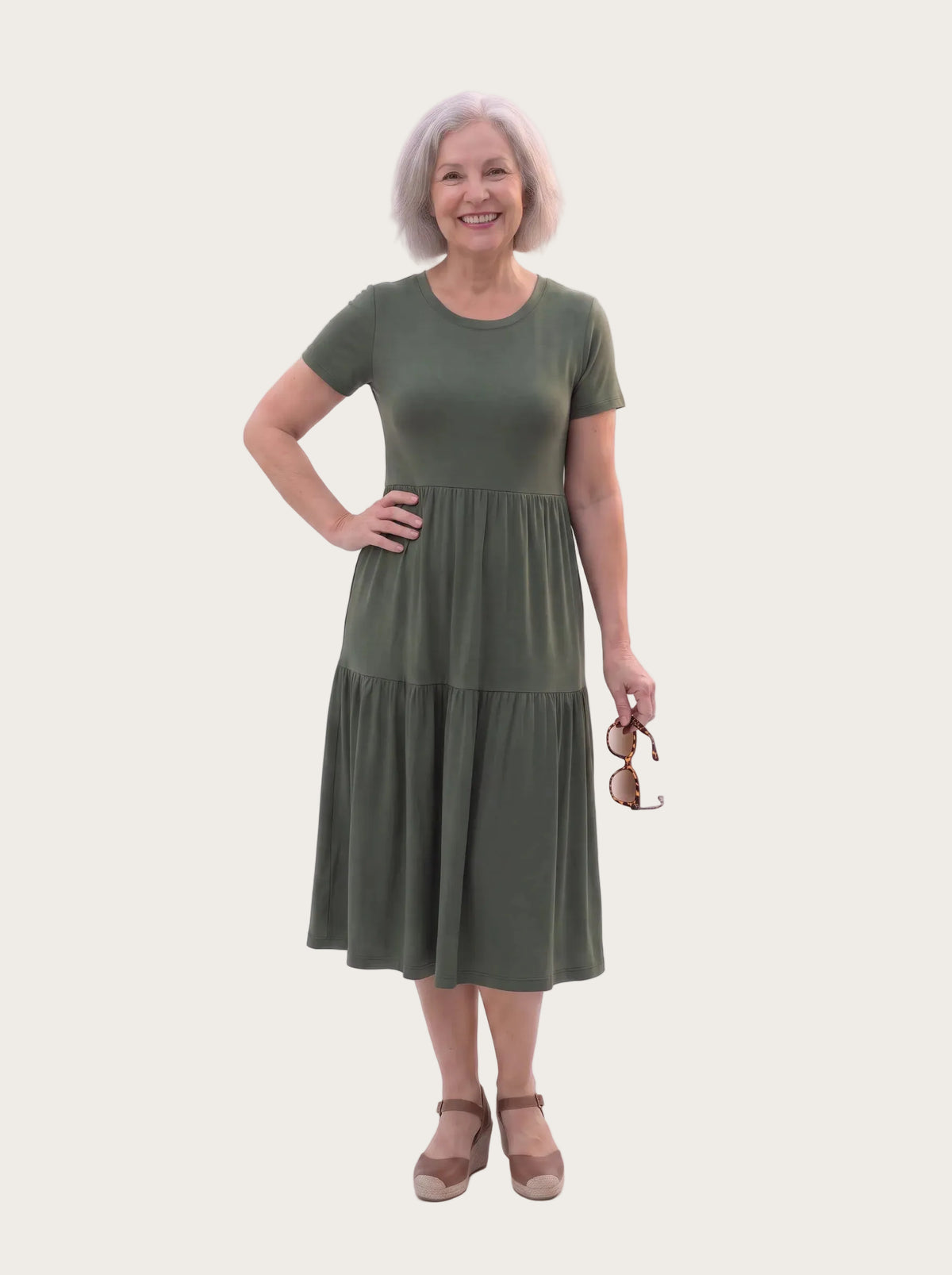 An older woman with short gray hair stands against a plain white studio background wearing a short-sleeve olive green midi dress with a gathered waist and holding a pair of sunglasses.