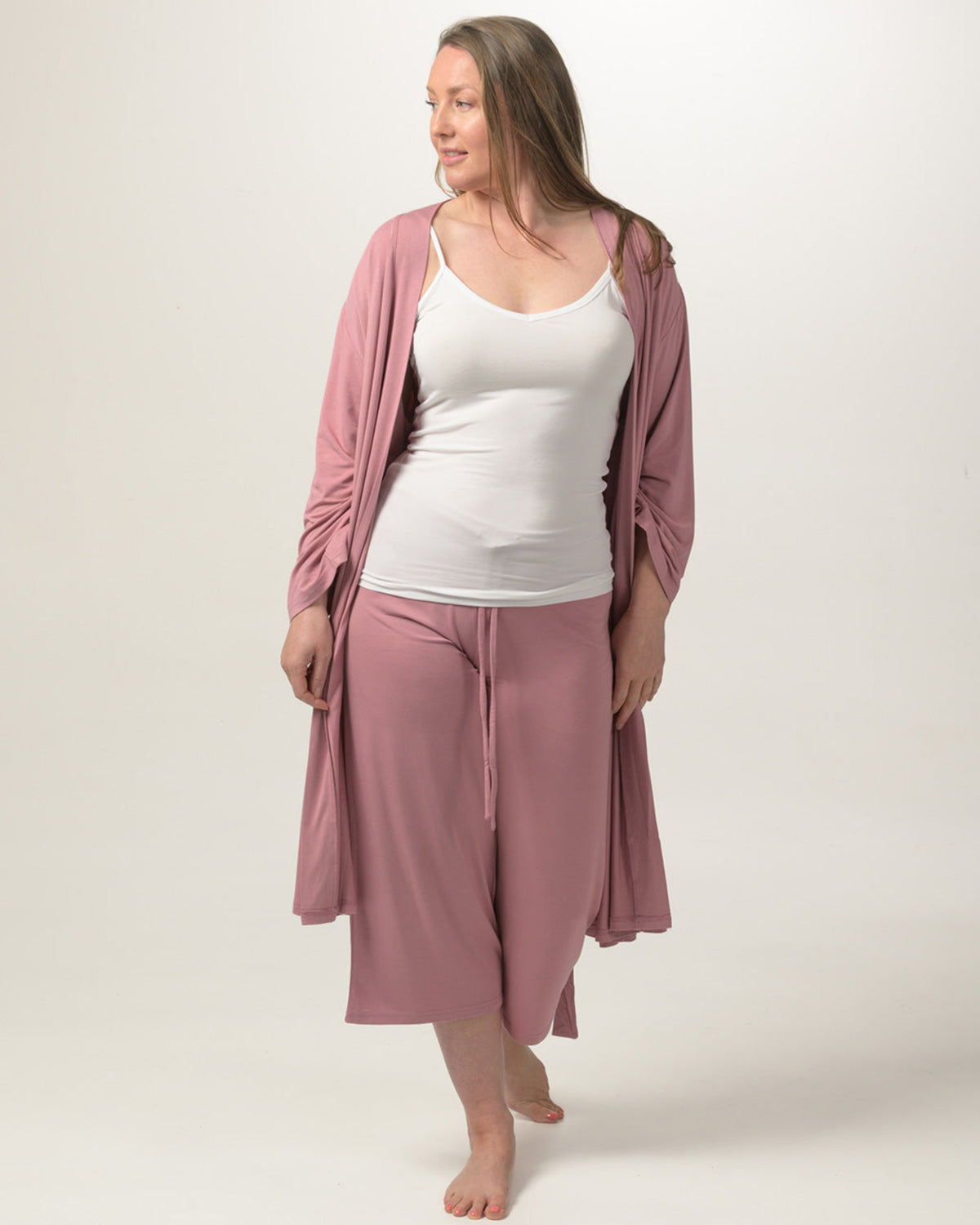 A woman modeling a matching bamboo  dusty rose lounge set consisting of a long-sleeve draped cardigan and high-waisted cropped pants, worn over a white tank top, against a plain light beige studio background.