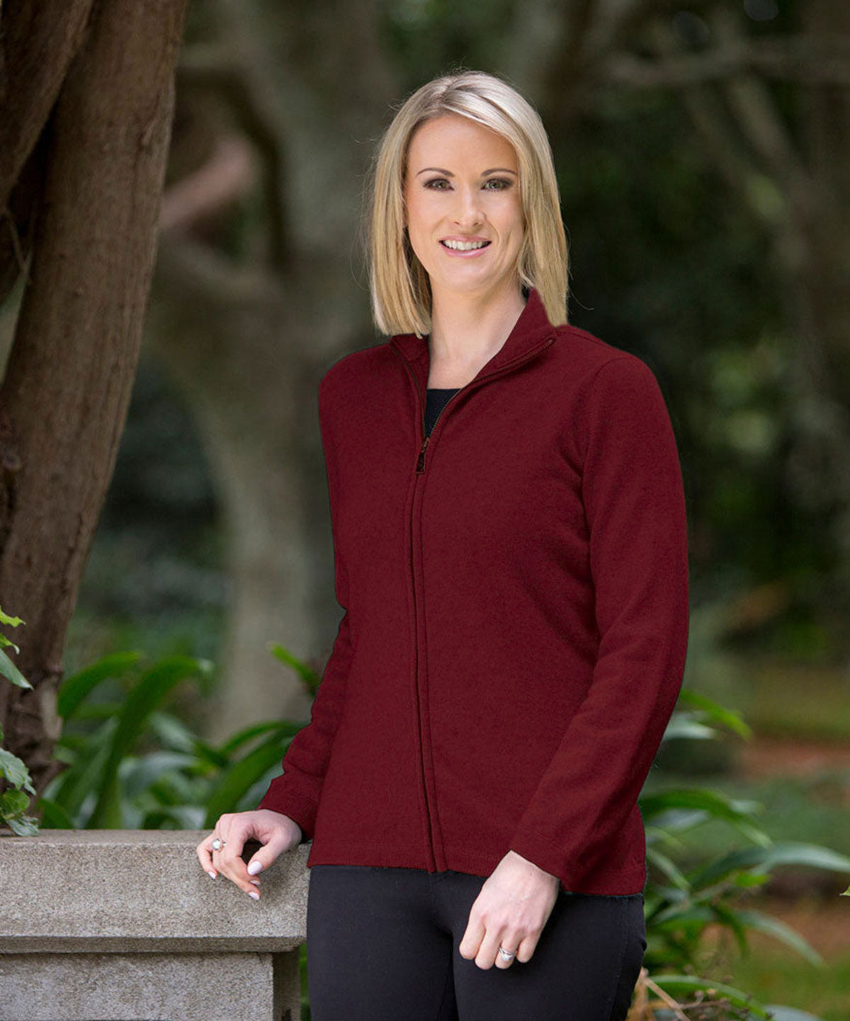 Woman wearing a possum merino  red jacket standing outdoors with greenery in the background