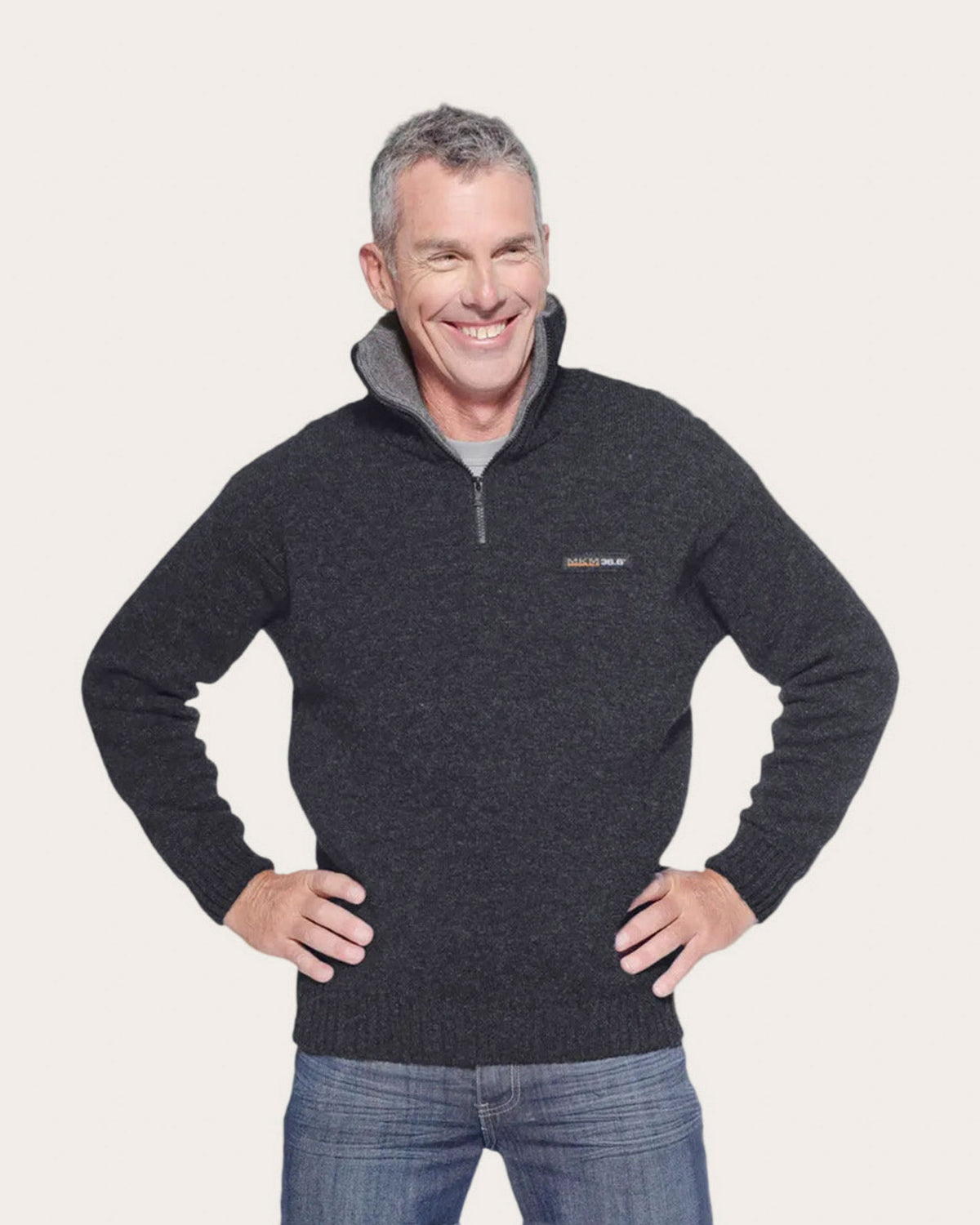 A smiling man wearing a dark charcoal gray quarter-zip possum merino pullover sweater with a gray inner collar and a small embroidered logo on the left chest, paired with blue jeans, against a solid off-white background.