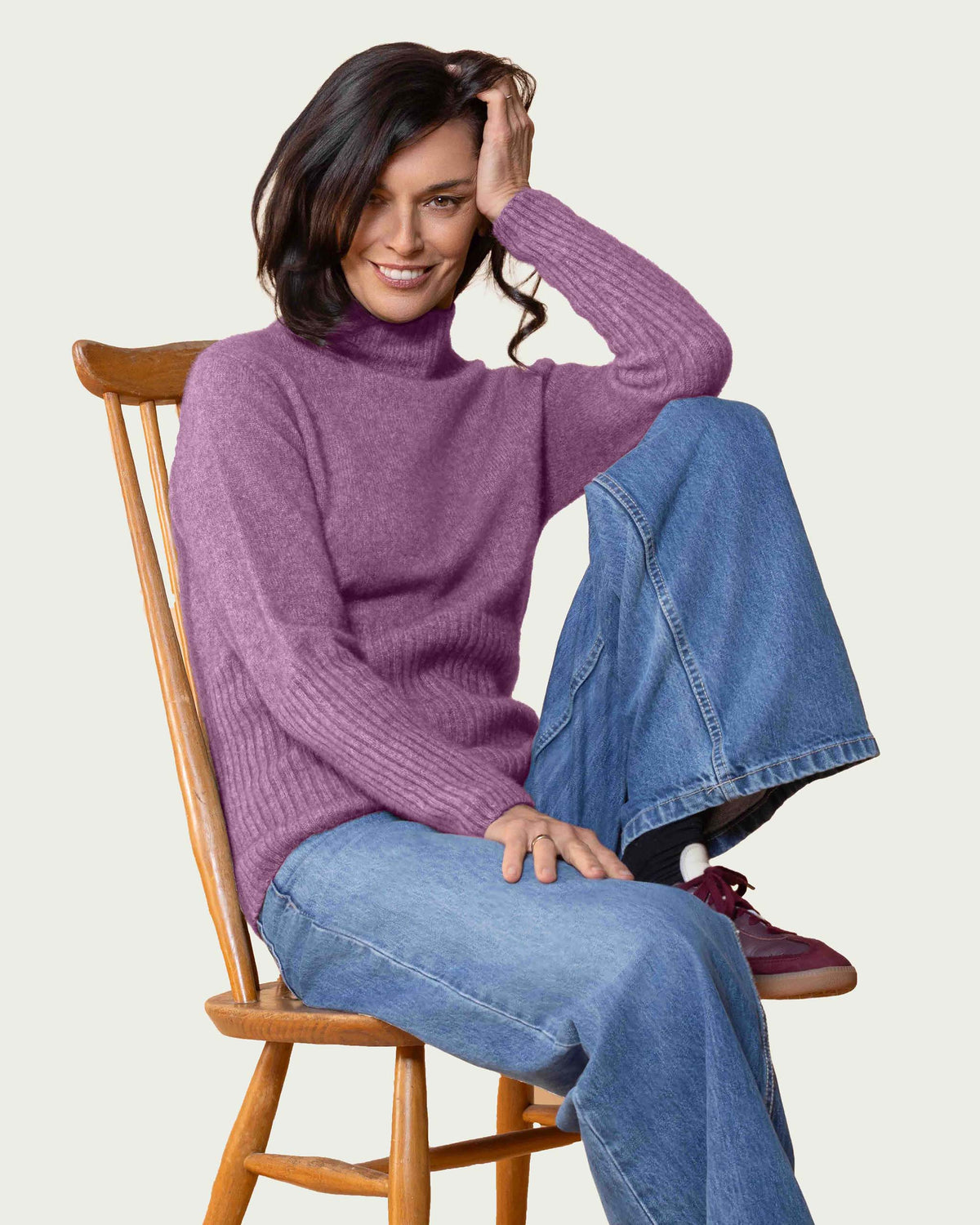 A woman wearing a ribbed dusky mauve possum merino turtleneck sweater and wide-leg blue jeans sits on a wooden chair against a light gray background.