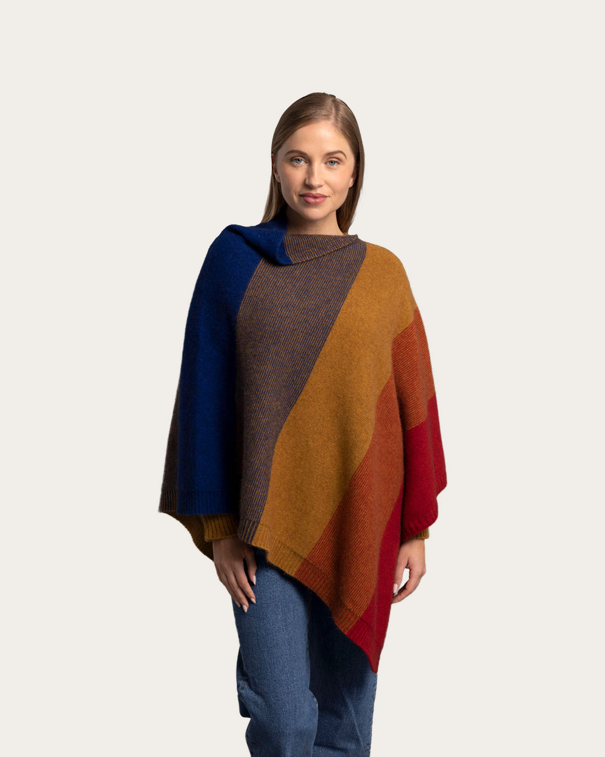 A woman modeling a multi-colored Possum Merino knit poncho with diagonal color-blocked panels in navy blue, brown, mustard yellow, orange, and red, paired with blue jeans against a light gray background.
