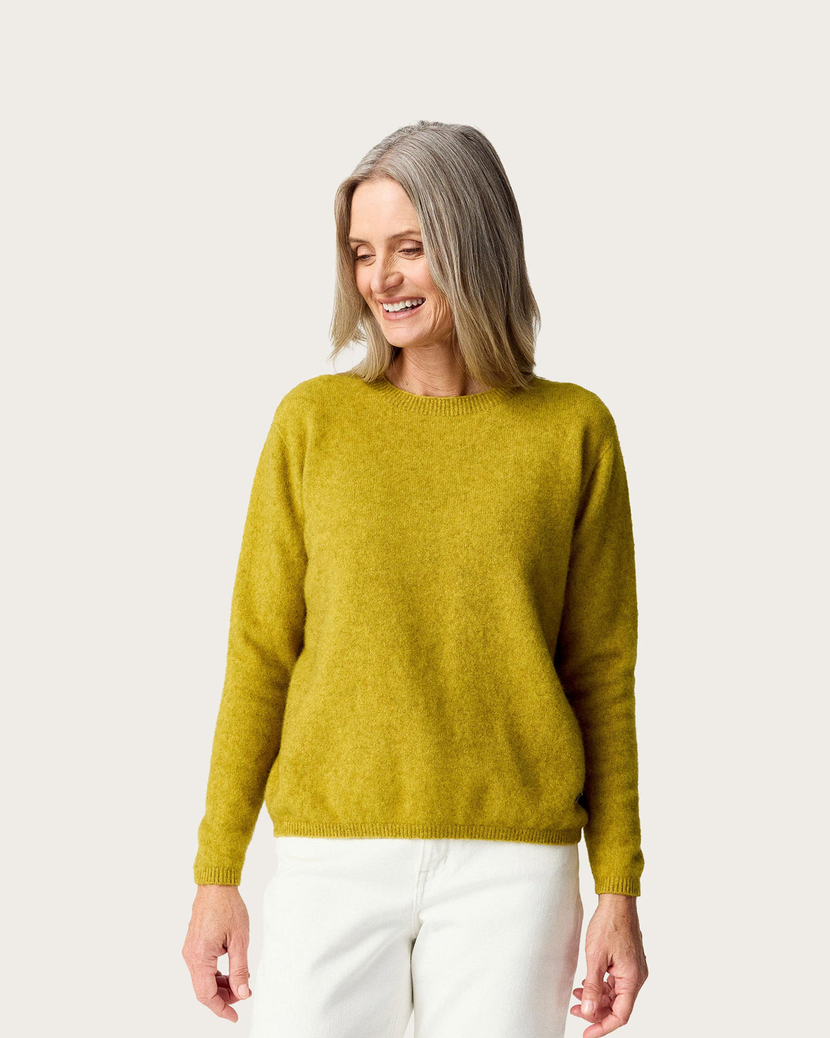 A woman with shoulder-length gray hair wearing a possum merino, textured mustard yellow sweater and white pants, standing against a solid light gray background.