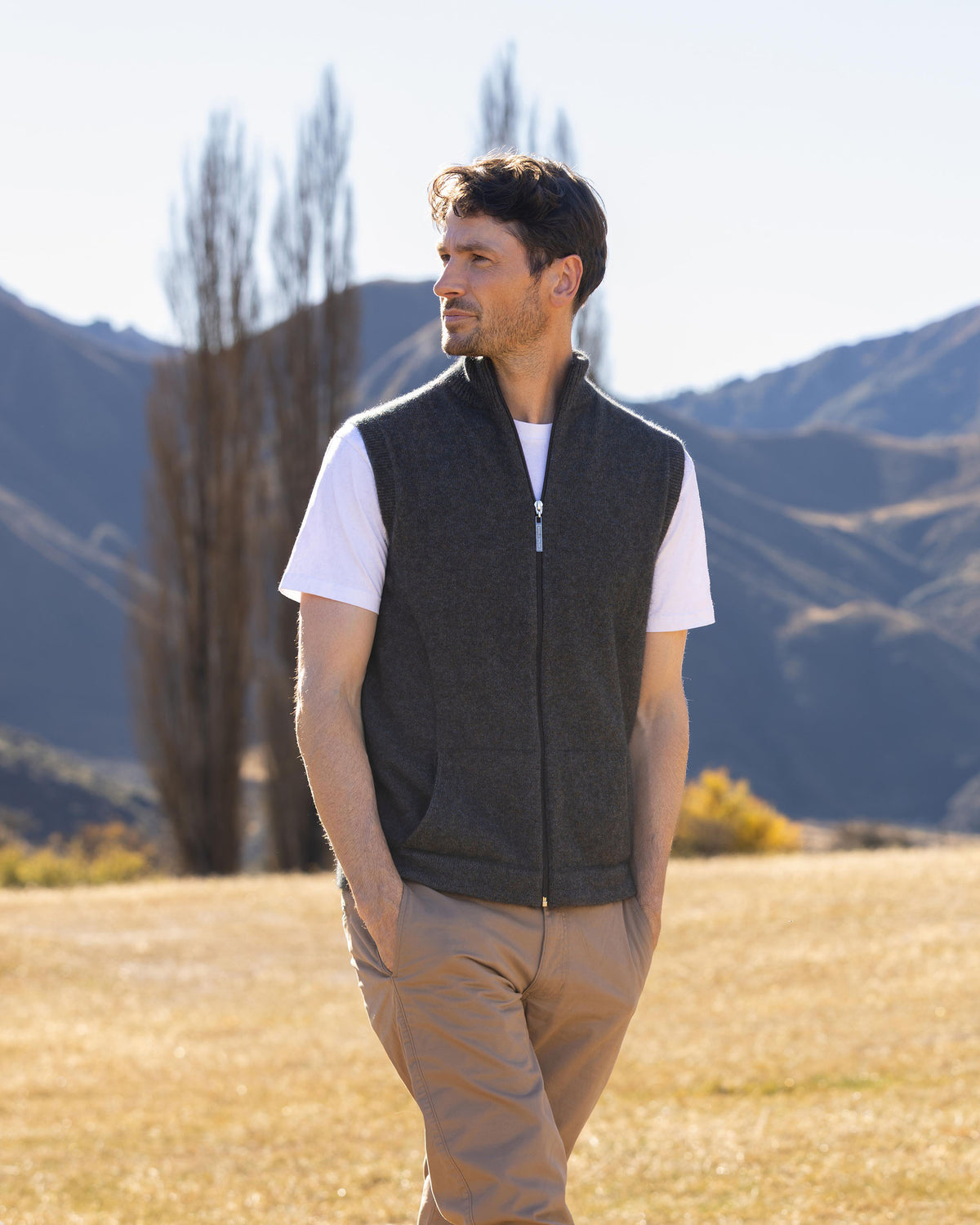 possum merino A man wearing a dark gray zip-up fleece vest over a white t-shirt and khaki pants, standing in a dry grassy field with mountains and trees in the background.