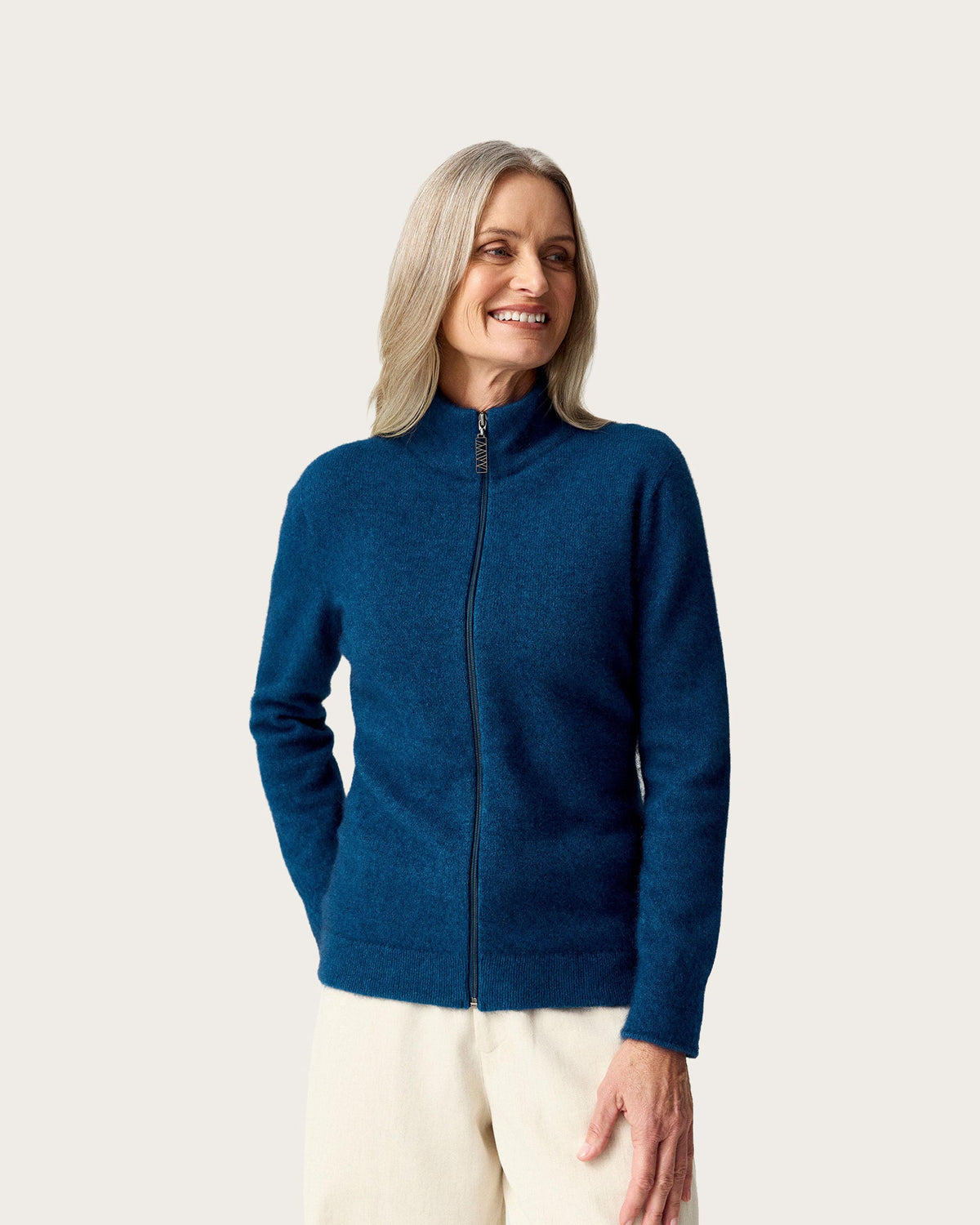 A smiling woman with blonde hair wearing a dark blue zip-up possum merino jacket and cream pants, standing against a solid light gray background.