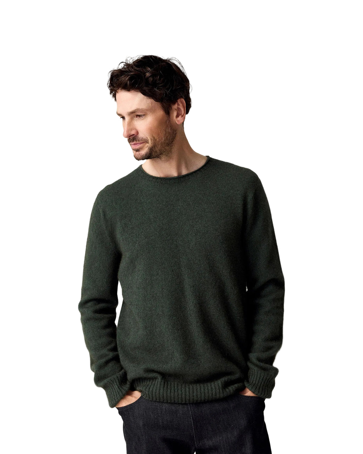 A man wearing a dark forest green crewneck possum merino knit sweater and dark jeans, standing against a solid light beige background.
