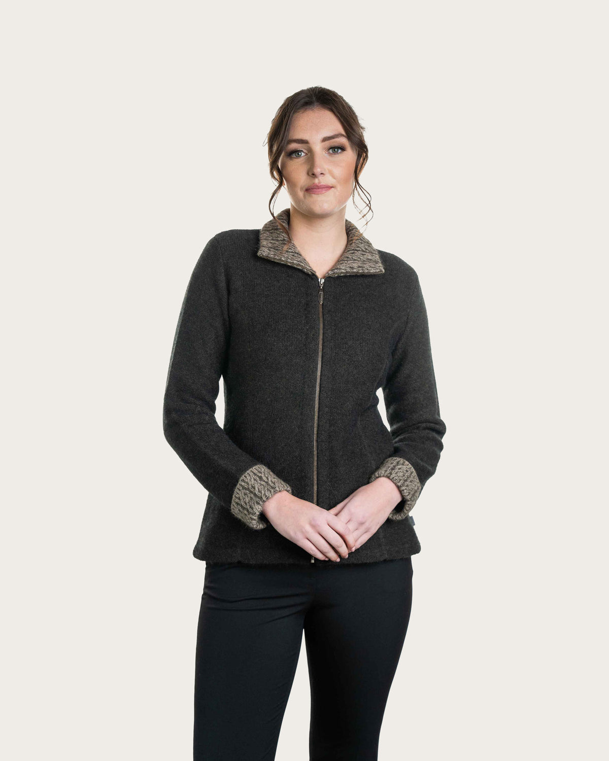 A woman modeling a dark expresso brown, possum merino, zip-up fleece jacket with textured beige and gray knit trim on the collar and cuffs, paired with black pants, against a solid light beige background.