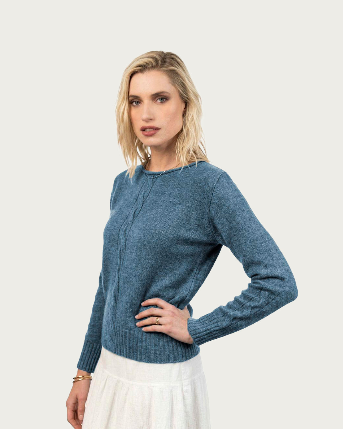 A blonde woman wearing a heathered slate blue possum merino knit sweater with a crew neck and ribbed cuffs, paired with a white skirt, photographed against a solid light gray background.