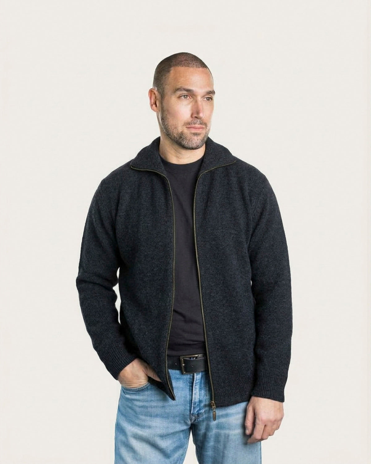 A man wearing a dark charcoal zip-up possum merino jacket over a black t-shirt and blue jeans, standing against a plain off-white background.
