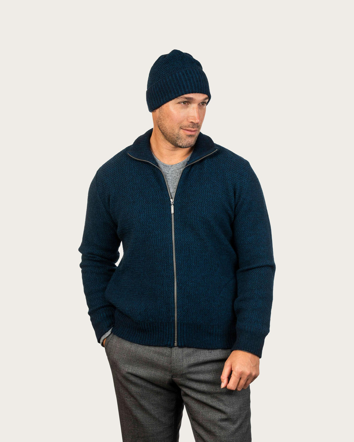 A man wearing a deep teal, possum merino zip-up sweater, a matching navy beanie, a gray t-shirt, and gray trousers, standing against a light gray studio background.