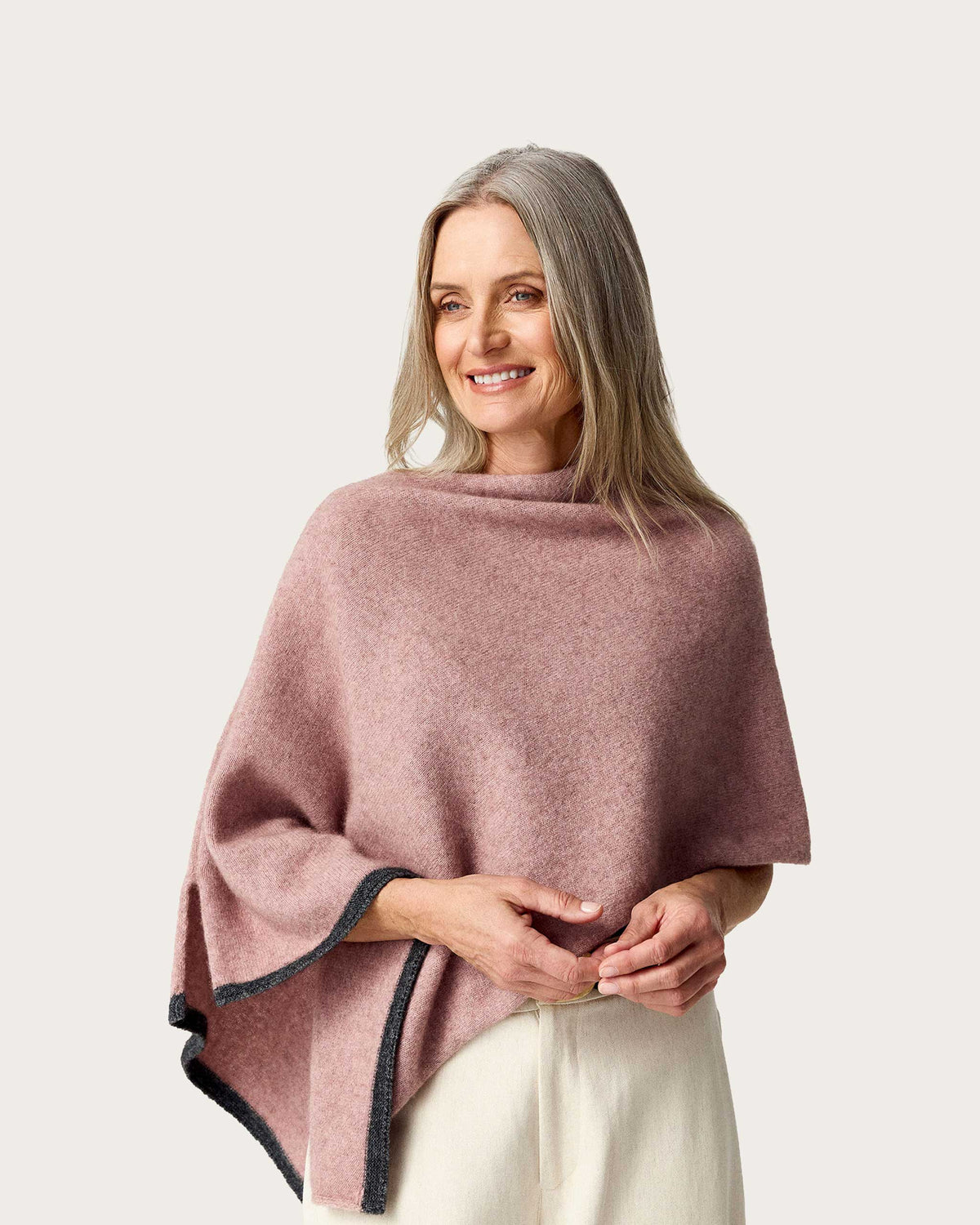 A woman wearing a dusty rose pink possum merino knit poncho with dark gray trim, paired with cream-colored pants, against a solid light gray background.