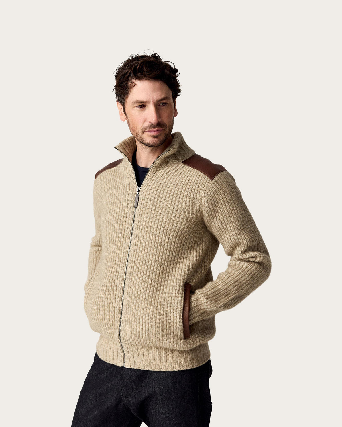 A man wearing a beige ribbed zip-up possum merino sweater with brown shoulder and pocket accents, paired with dark jeans, against a light gray studio background.