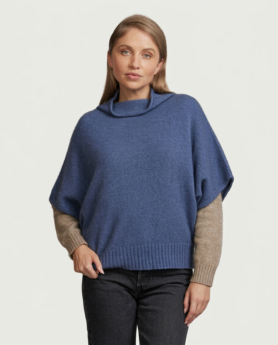 A woman wearing a blue possum merino knit poncho sweater with beige ribbed sleeves, paired with dark jeans, against a light gray studio background.