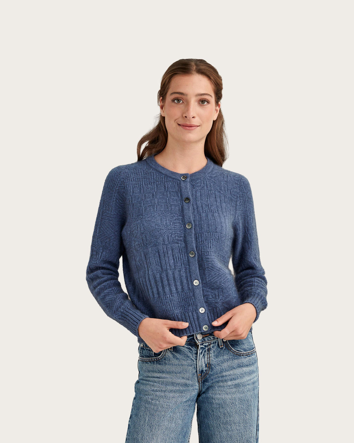 A woman wearing a blue textured possum merino knit cardigan with a button-up front and blue jeans, standing against a solid light beige background.