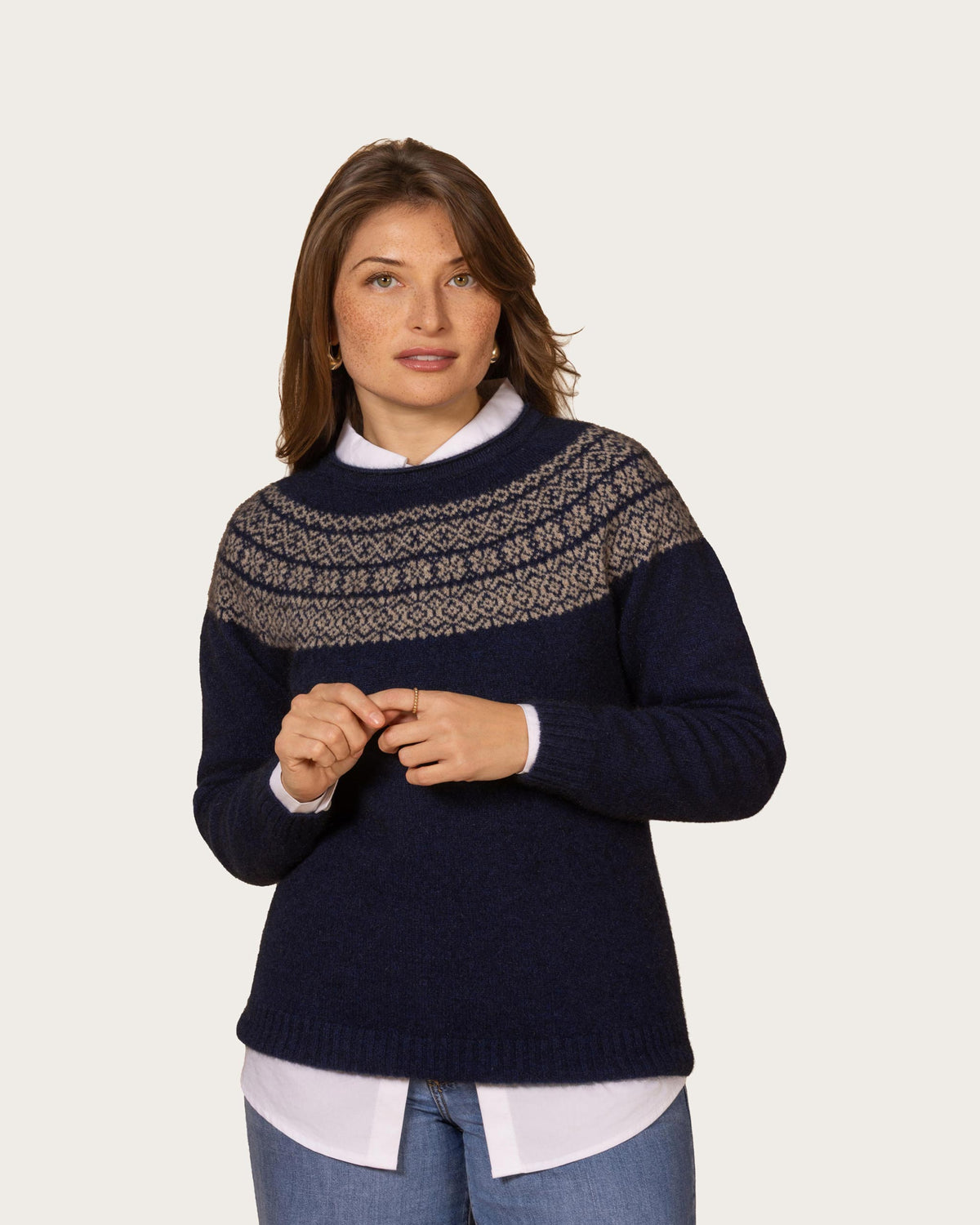 A woman wearing a navy blue possum merino knit sweater with a beige and gray Fair Isle yoke pattern, styled over a white collared shirt and blue jeans, against a light gray studio background.