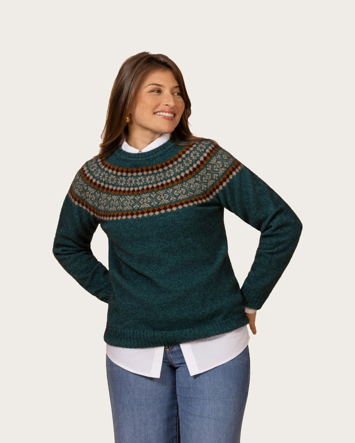 A woman modeling a dark teal possum merino knit sweater with a brown, cream, and gray Fair Isle yoke pattern, styled over a white collared shirt and blue jeans against a light beige background.