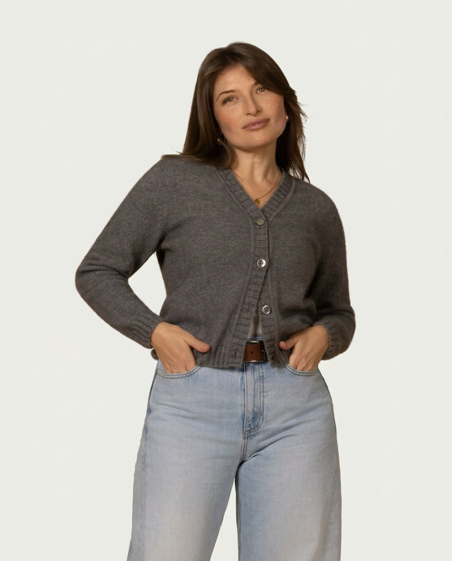 A woman modeling a dark gray button-up possum merino cardigan sweater with light-wash blue jeans against a plain light gray studio background.