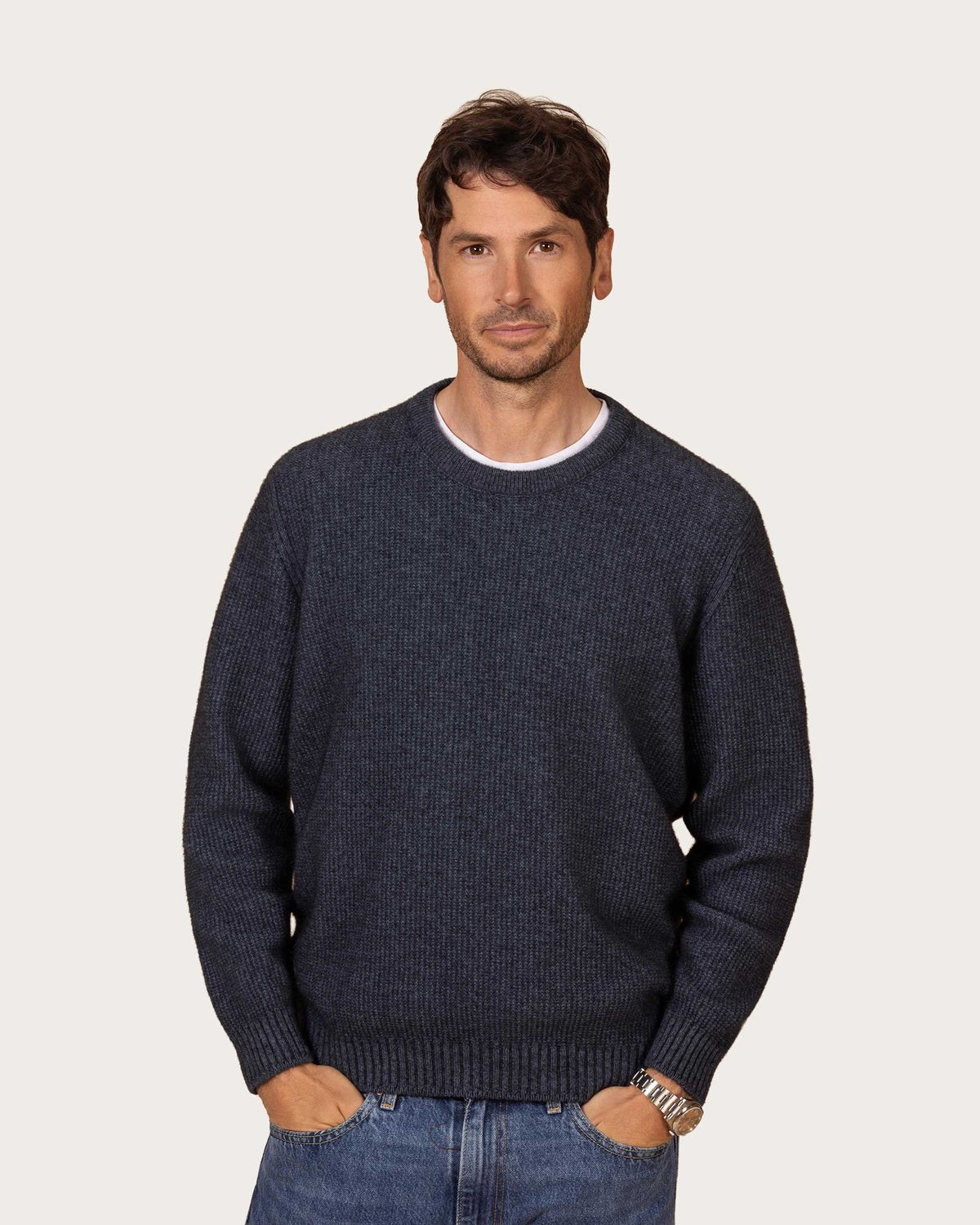 A man wearing a dark denim blue textured possum merino knit crewneck sweater over a white undershirt and blue jeans, standing against a solid light beige background.