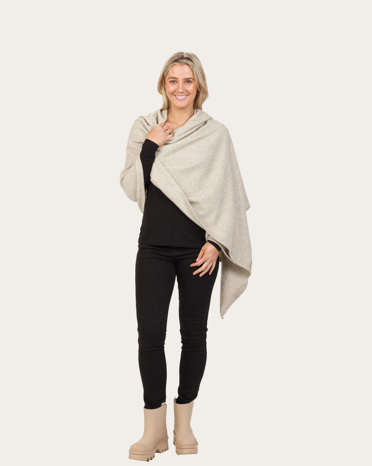 A woman modeling a light beige possum merino knit wrap or poncho draped over her shoulders, paired with a black long-sleeve top, black skinny pants, and beige ankle boots, against a solid off-white background.