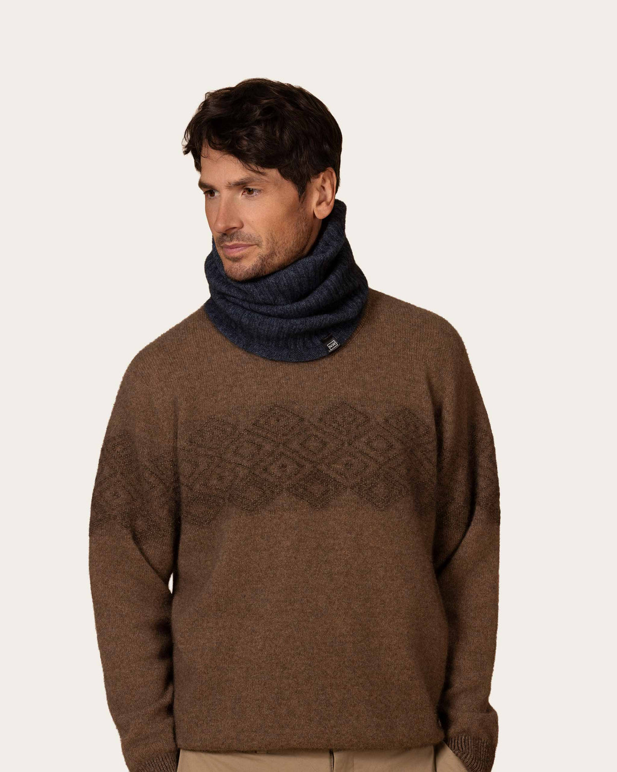 A man wearing a brown knit sweater with a dark geometric pattern across the chest and a denim blue possum merino ribbed neck warmer, photographed against a plain light beige background.