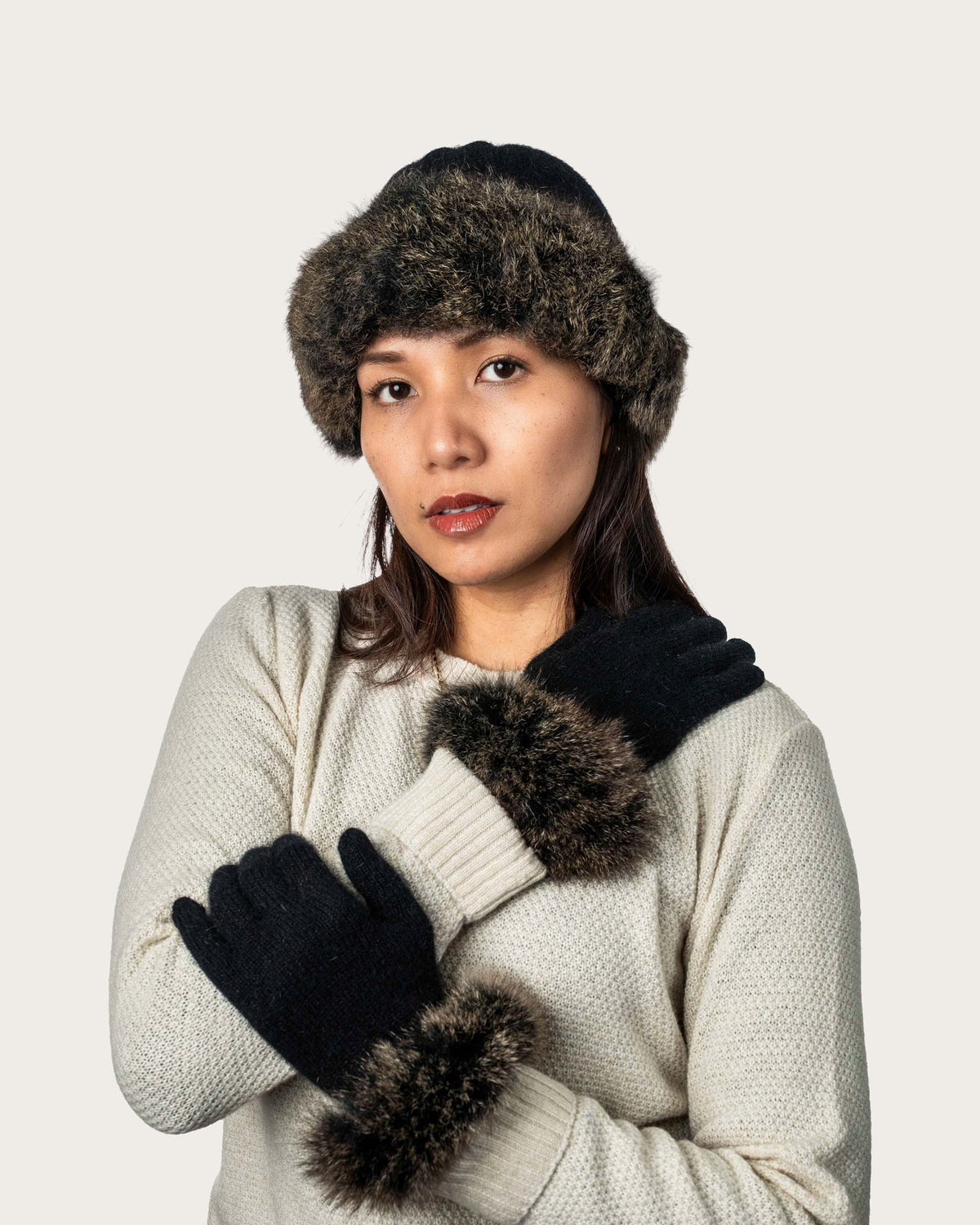 A woman wearing a dark brown and black fur-trimmed possum merino beanie, black knit gloves with matching fur trim, and a cream-colored knit sweater, posed against a solid light gray background.