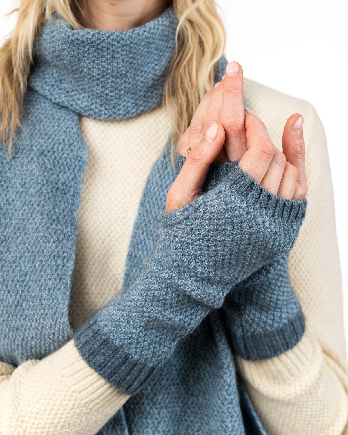 A close-up of a person wearing a chunky knit blue possum merino scarf and matching fingerless gloves over a cream-colored sweater, against a plain white background.