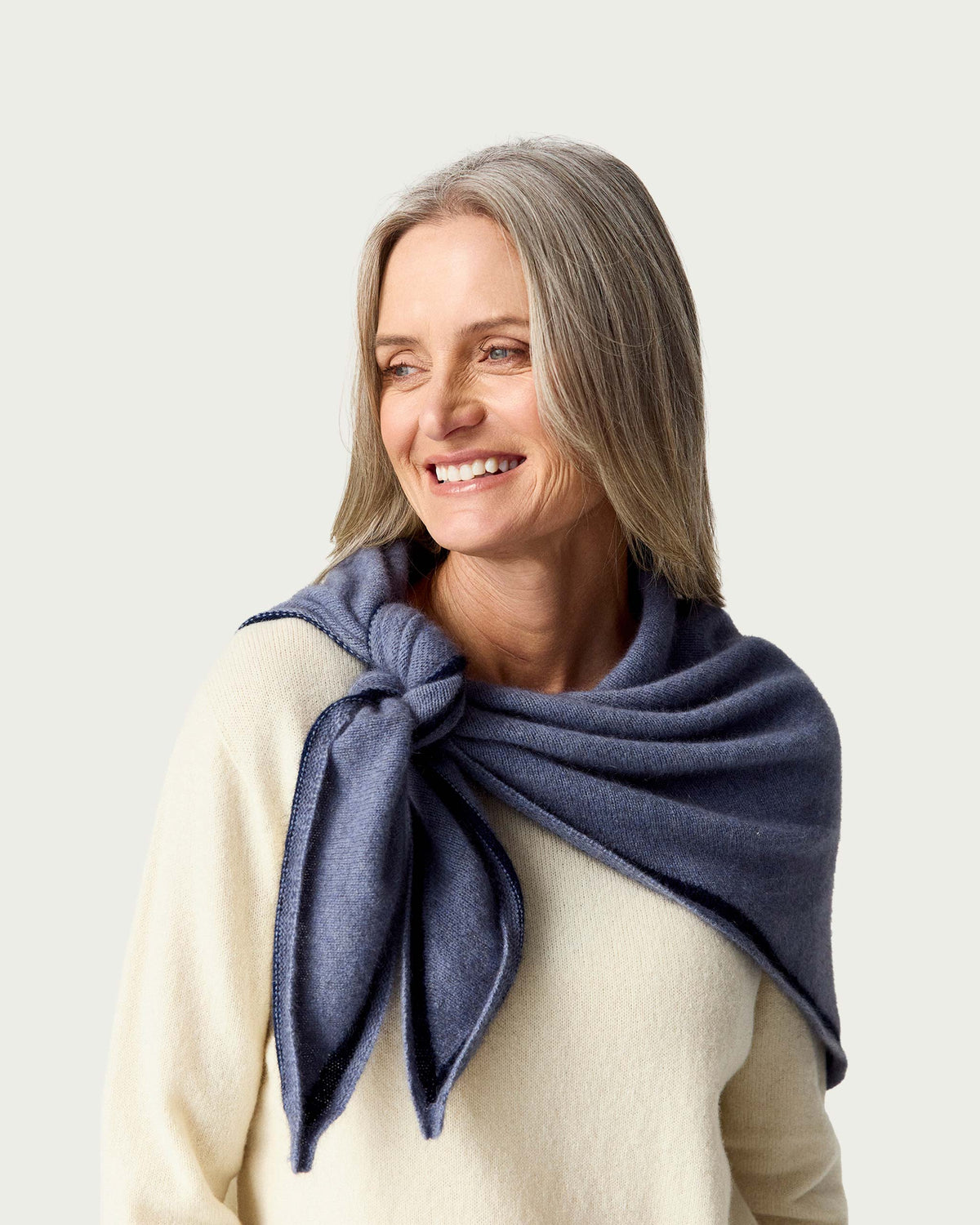 A smiling woman with gray hair models a navy blue possum merino knit scarf tied around her neck over a cream-colored sweater, set against a solid light gray background.