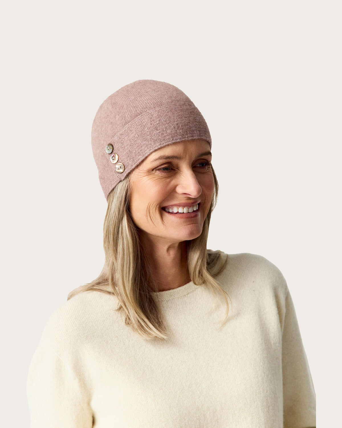 A smiling woman wearing a dusty rose possum merino  knit beanie with three decorative buttons on the side, paired with a cream-colored knit sweater, set against a solid light beige background.