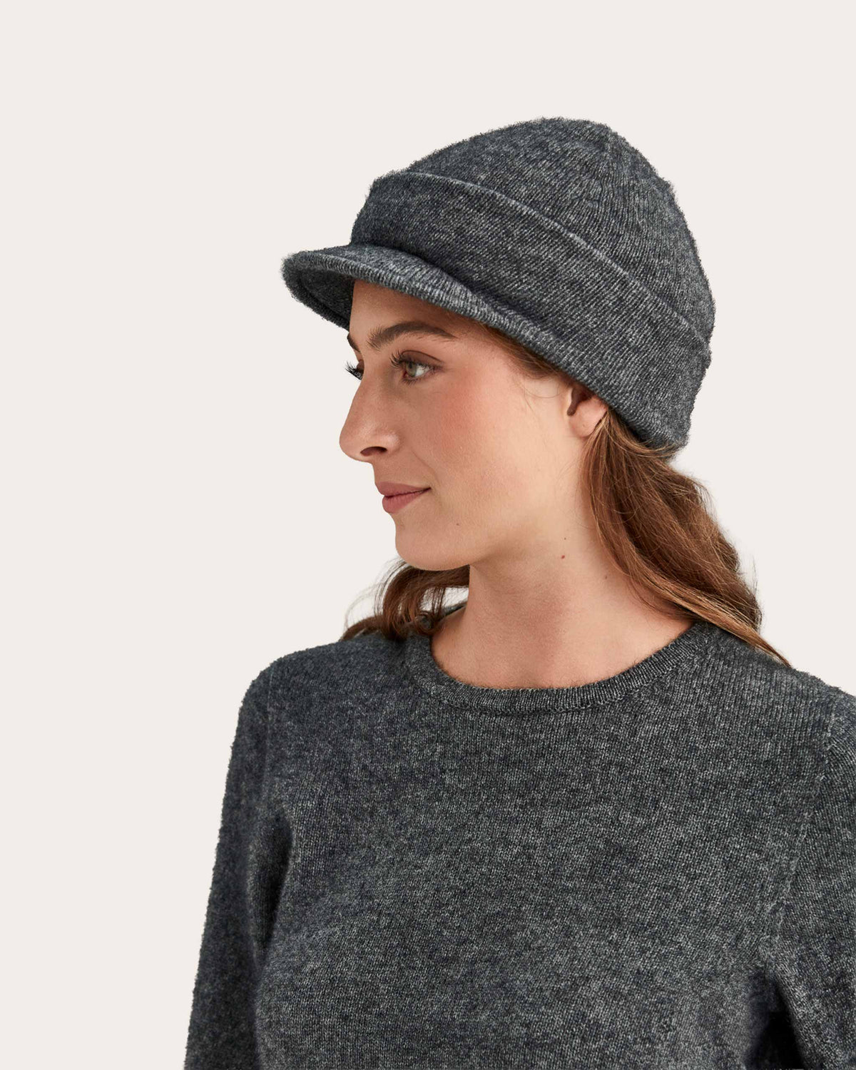 A woman in profile wearing a dark gray possum merino knit beanie with a small flat brim and a matching dark gray knit top, set against a plain light beige background.