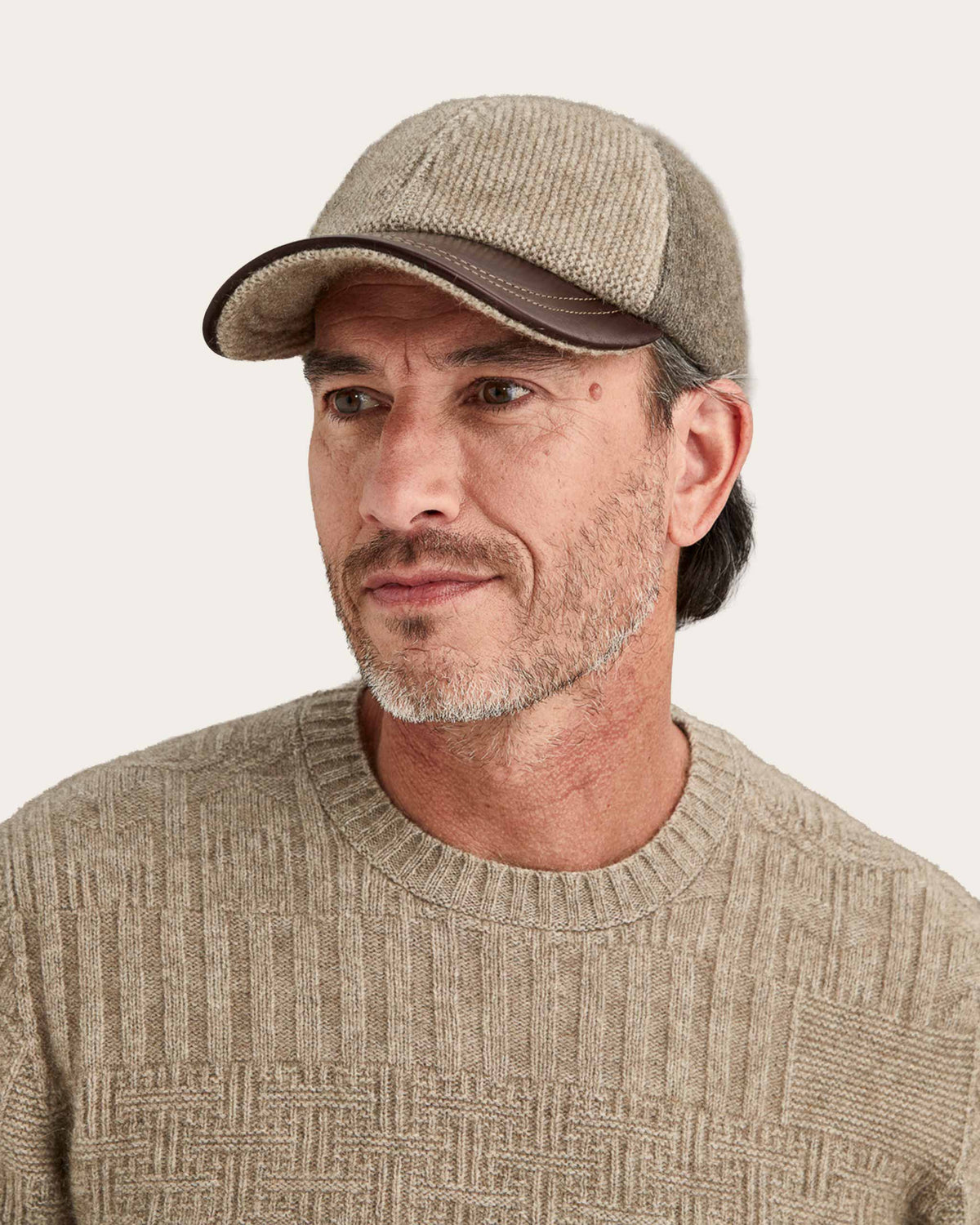 A man wearing a textured possum merino baseball cap in beige and brown, paired with a matching beige knit sweater, photographed against a solid off-white background.
