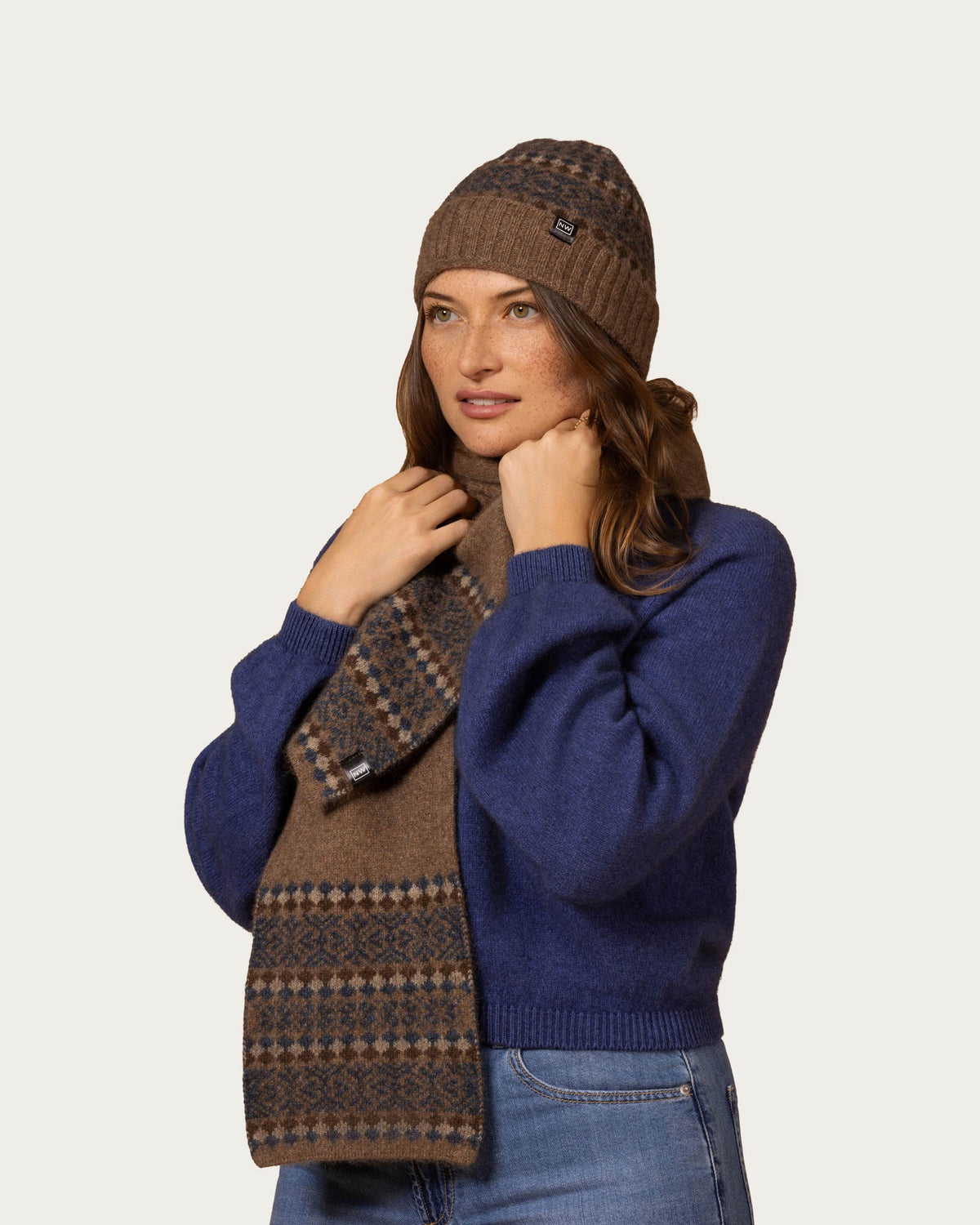 A woman wearing a brown possum merino knit beanie, a blue knit sweater, and a brown patterned scarf, posed against a solid light gray background.