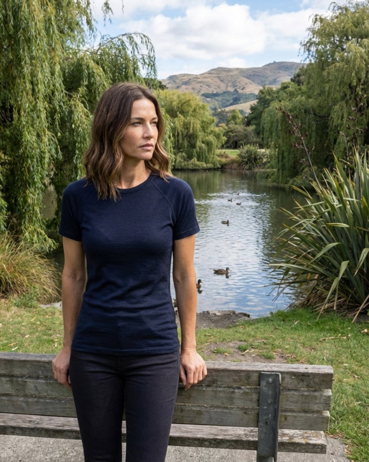 Woman in a merino  navy blue t-shirt standing by a pond with greenery and mountains in the background