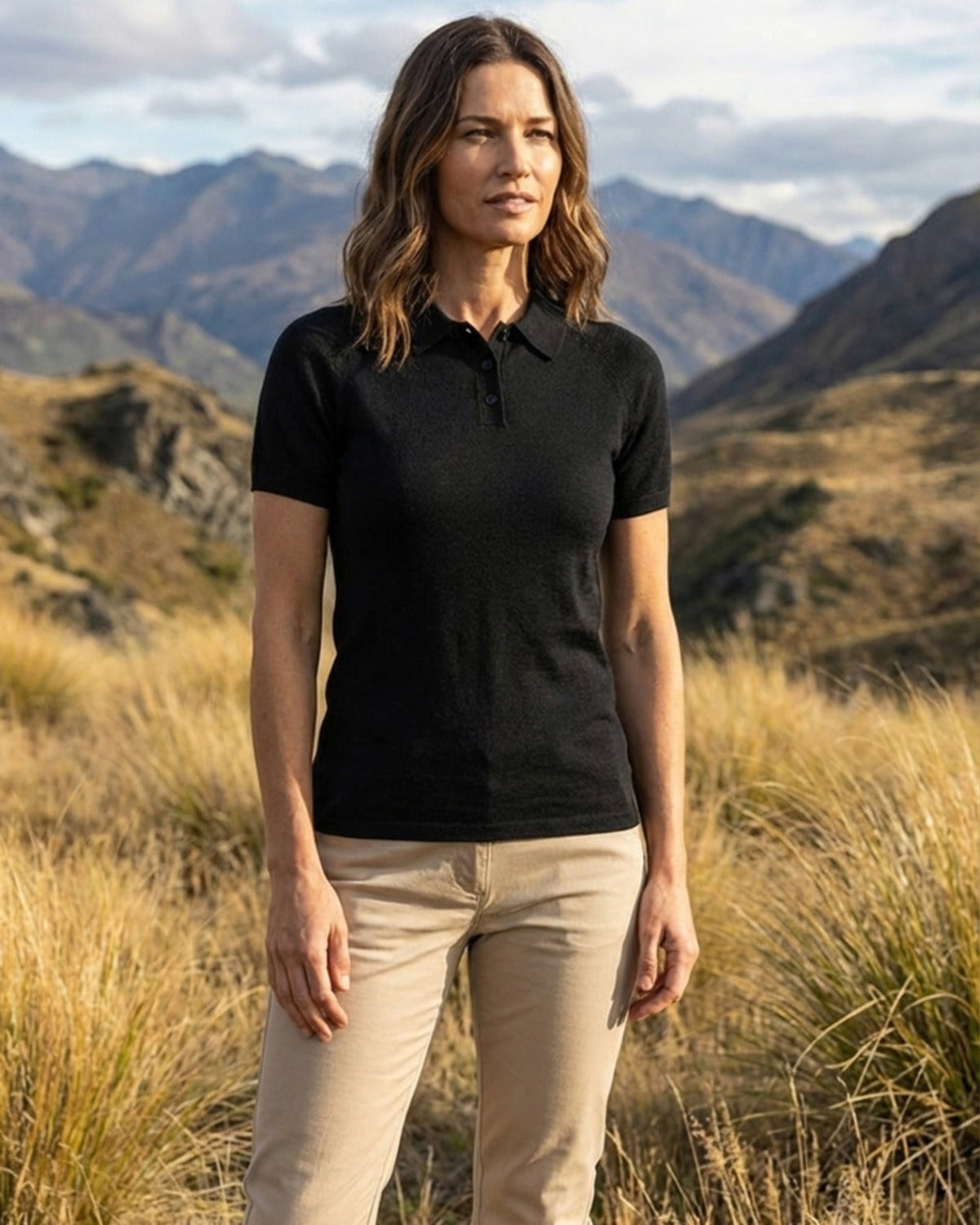 Woman wearing a merino black polo shirt and beige pants standing in a mountainous landscape.