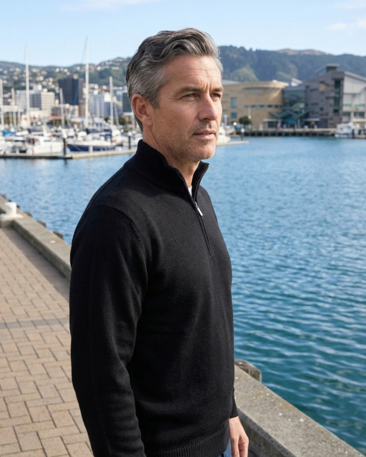 Man in a merino black sweater standing by a waterfront with buildings and mountains in the background
