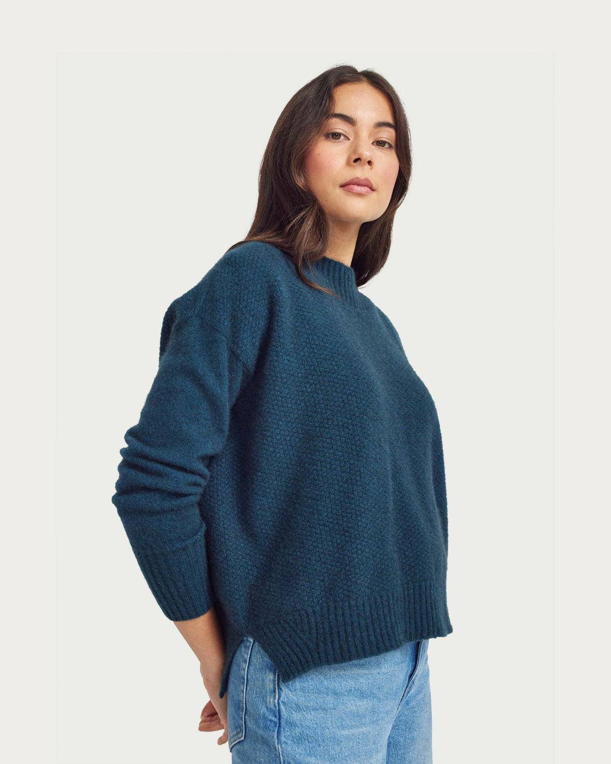 A woman wearing a dark teal textured knit possum merino sweater and light blue jeans, photographed against a solid off-white background.