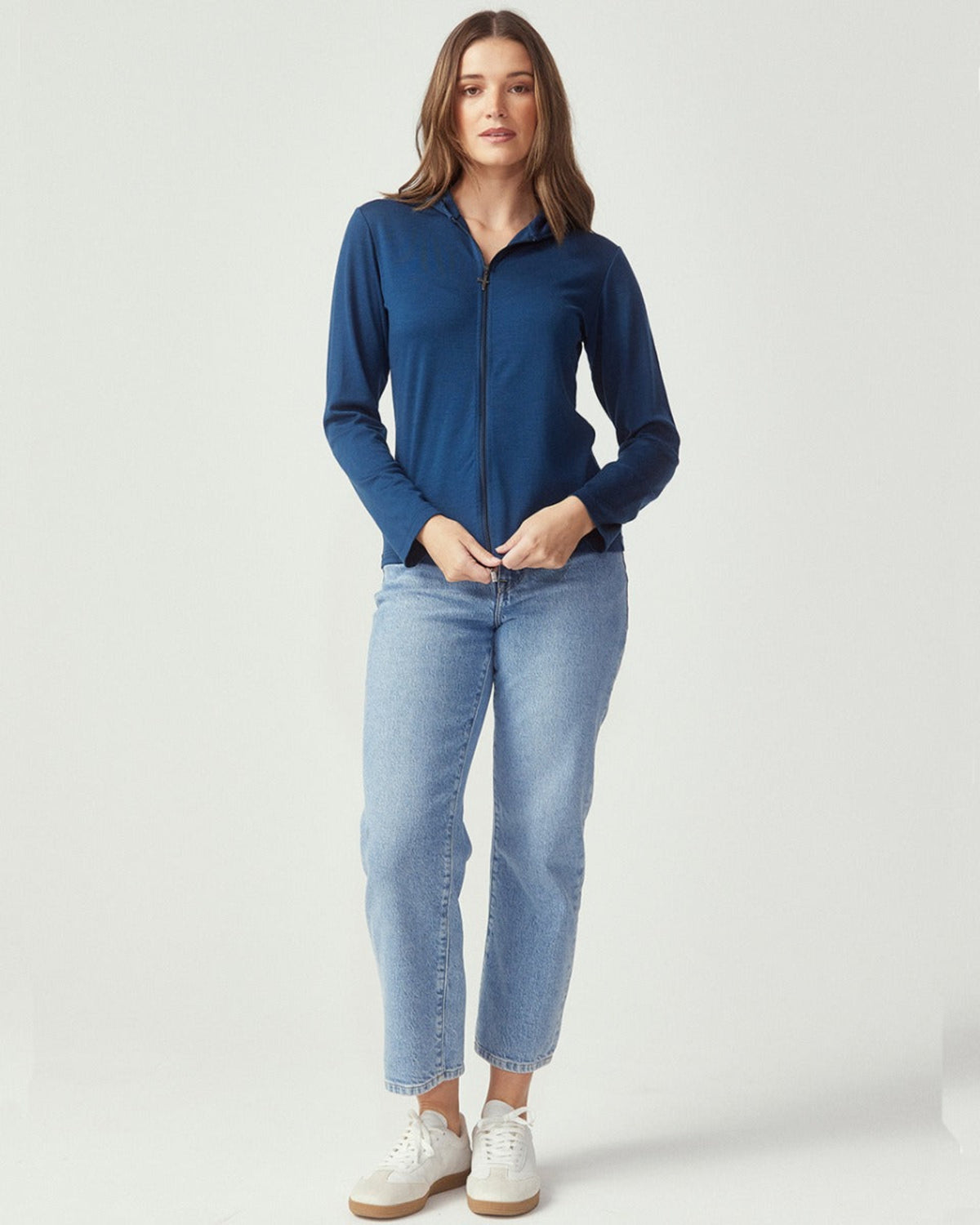 Woman wearing a merino blue zip-up top and light blue jeans on a white background