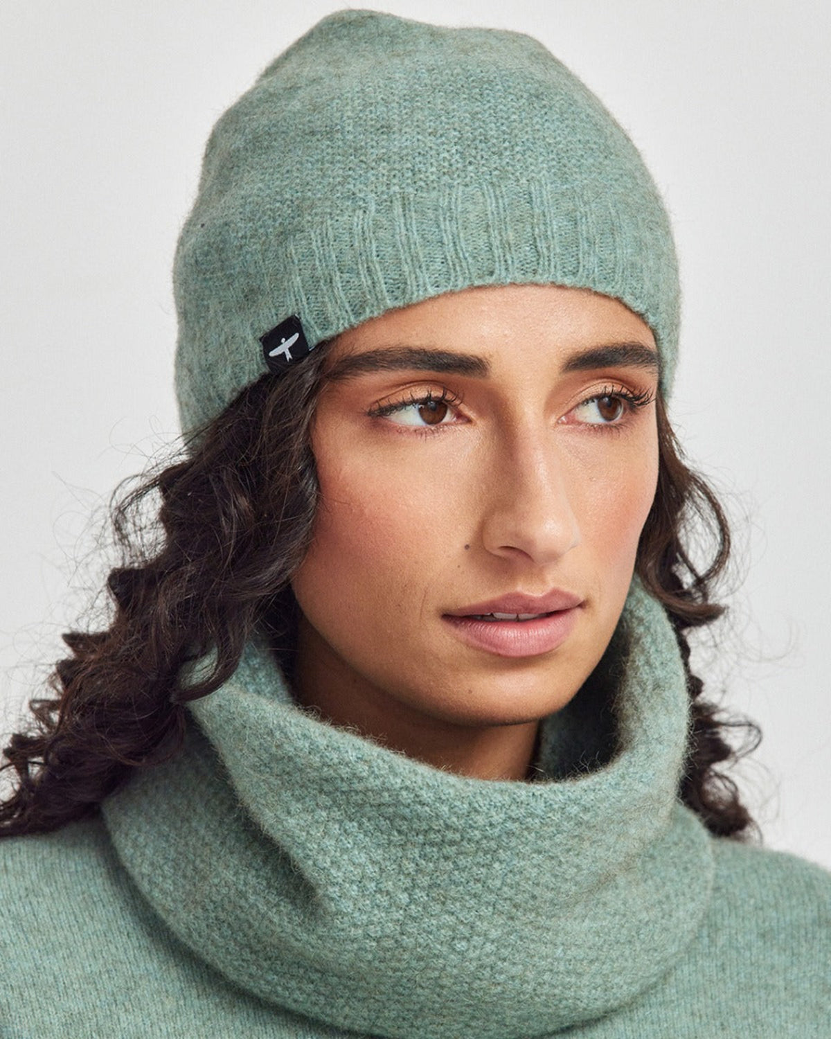 Possum merino Woman wearing a green knit beanie and scarf against a light gray background