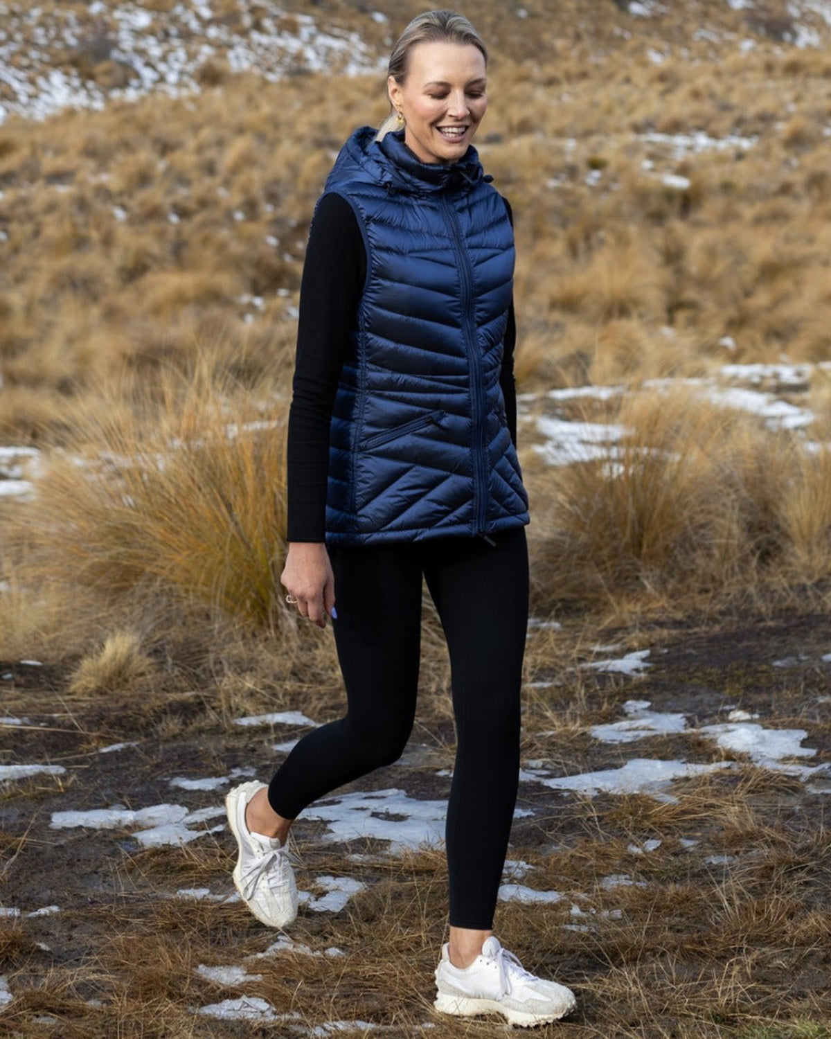 Woman wearing a blue puffer vest in a natural setting with dry grass and patches of snow.