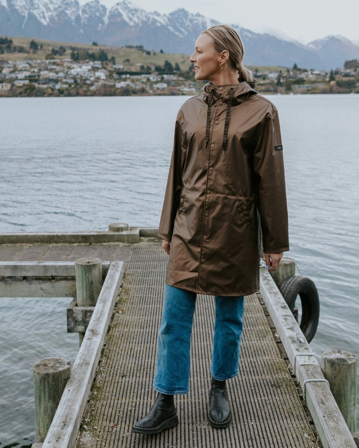 Person wearing a brown raincoat standing on a wooden dock by a body of water with mountains in the background.