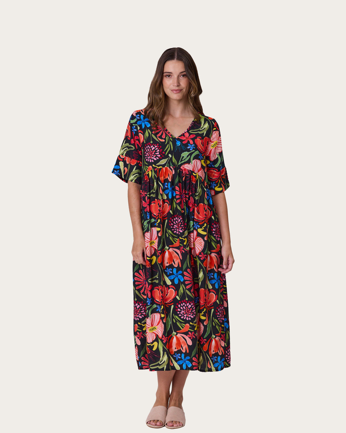 A woman modeling the Ella Dress by Democracy, a short-sleeve midi dress with a vibrant multicolor floral print on a black background, paired with neutral flat sandals, against a solid light beige backdrop.