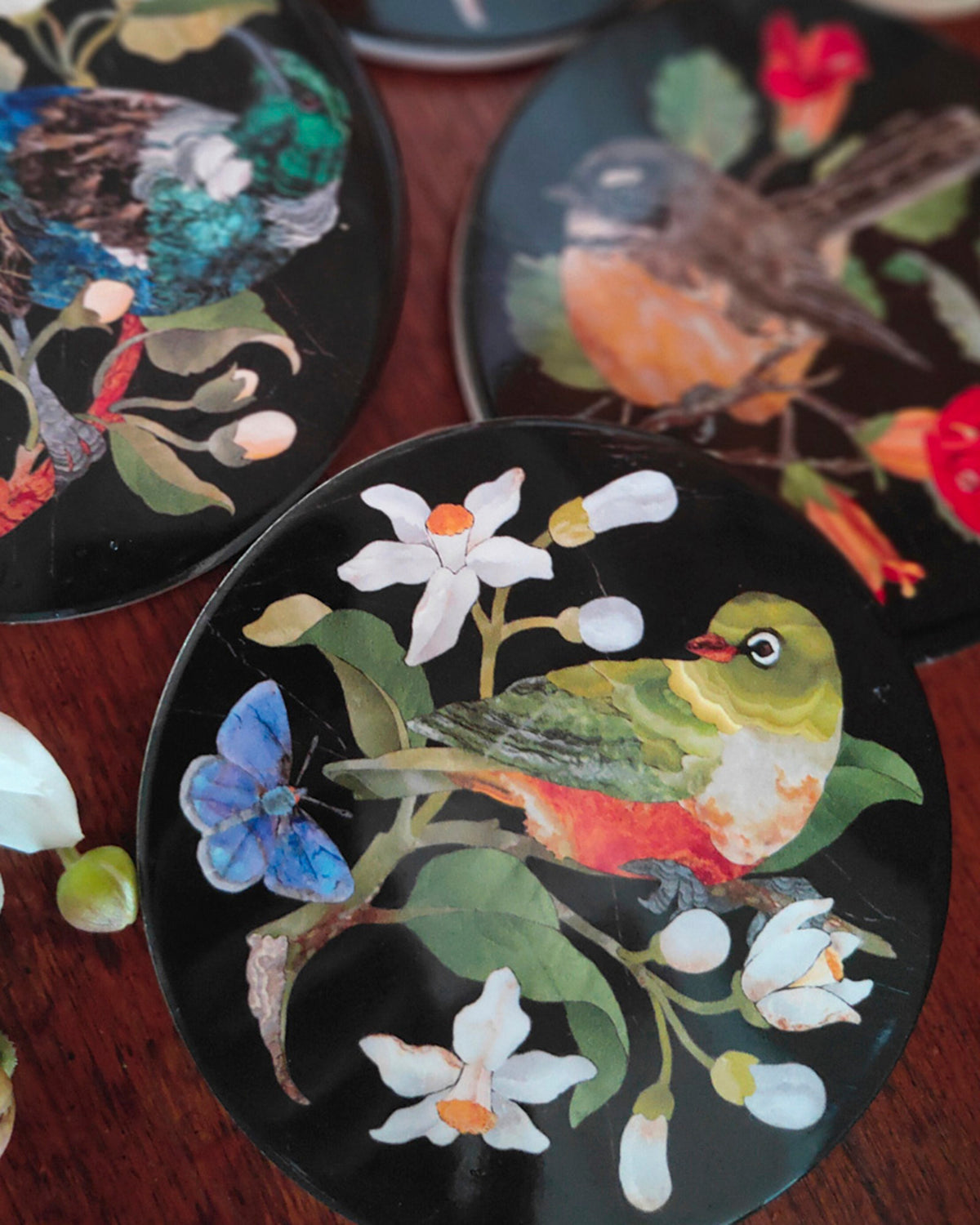 Three round black decorative coasters with intricate inlay artwork of birds, flowers, and butterflies, arranged on a dark wooden surface.