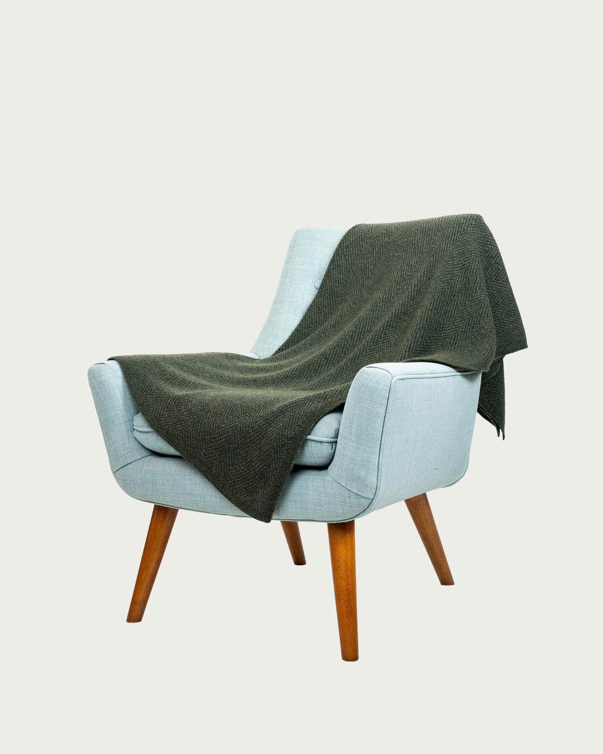 A light blue upholstered armchair with wooden legs, draped with a dark olive green possum merino knit throw blanket, set against a solid off-white background.