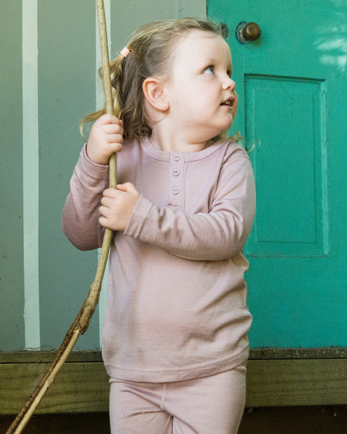 merino Child in pink outfit holding a stick, standing in front of a green door.