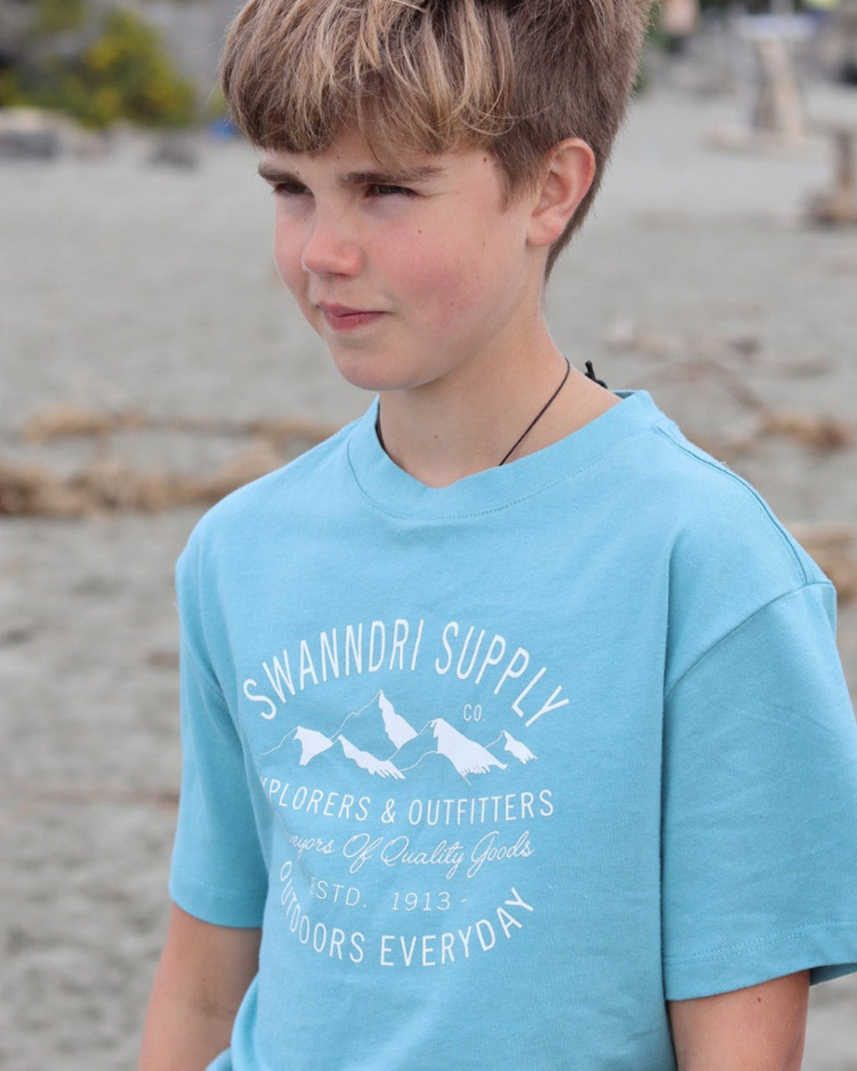 Cotton Young boy wearing a blue t-shirt with 'Swandri Supply' branding on a beach.