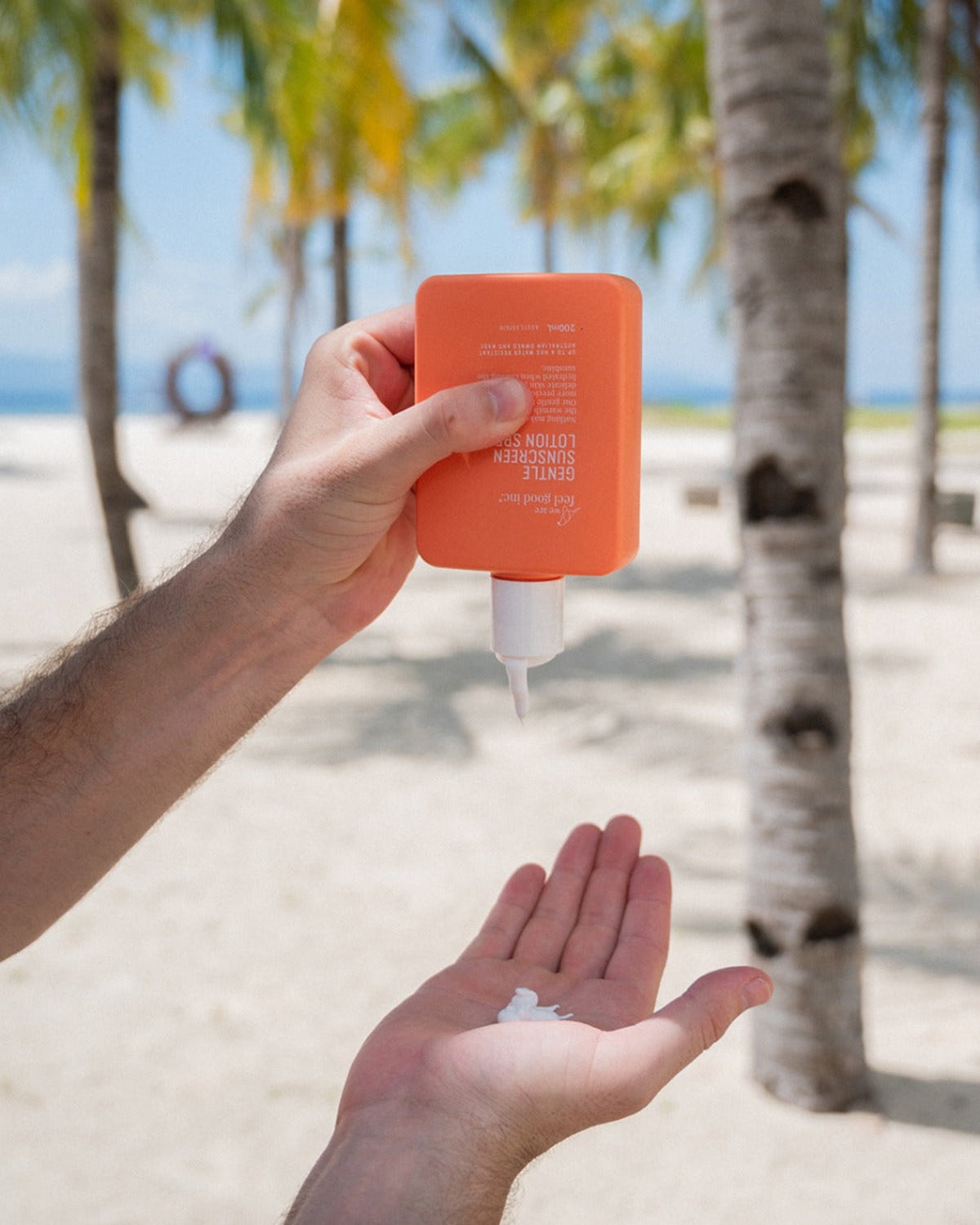 Person applying SPF50+ sunscreen lotion on a beach with palm trees in the background