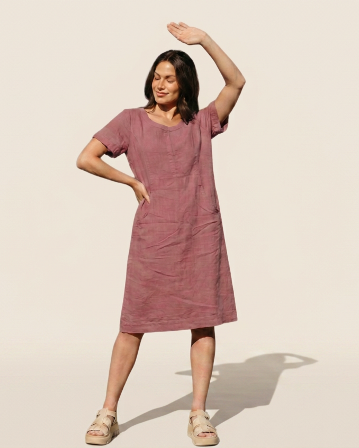 A woman modeling a dusty rose hemp dress with short sleeves and side pockets, standing against a plain beige background.